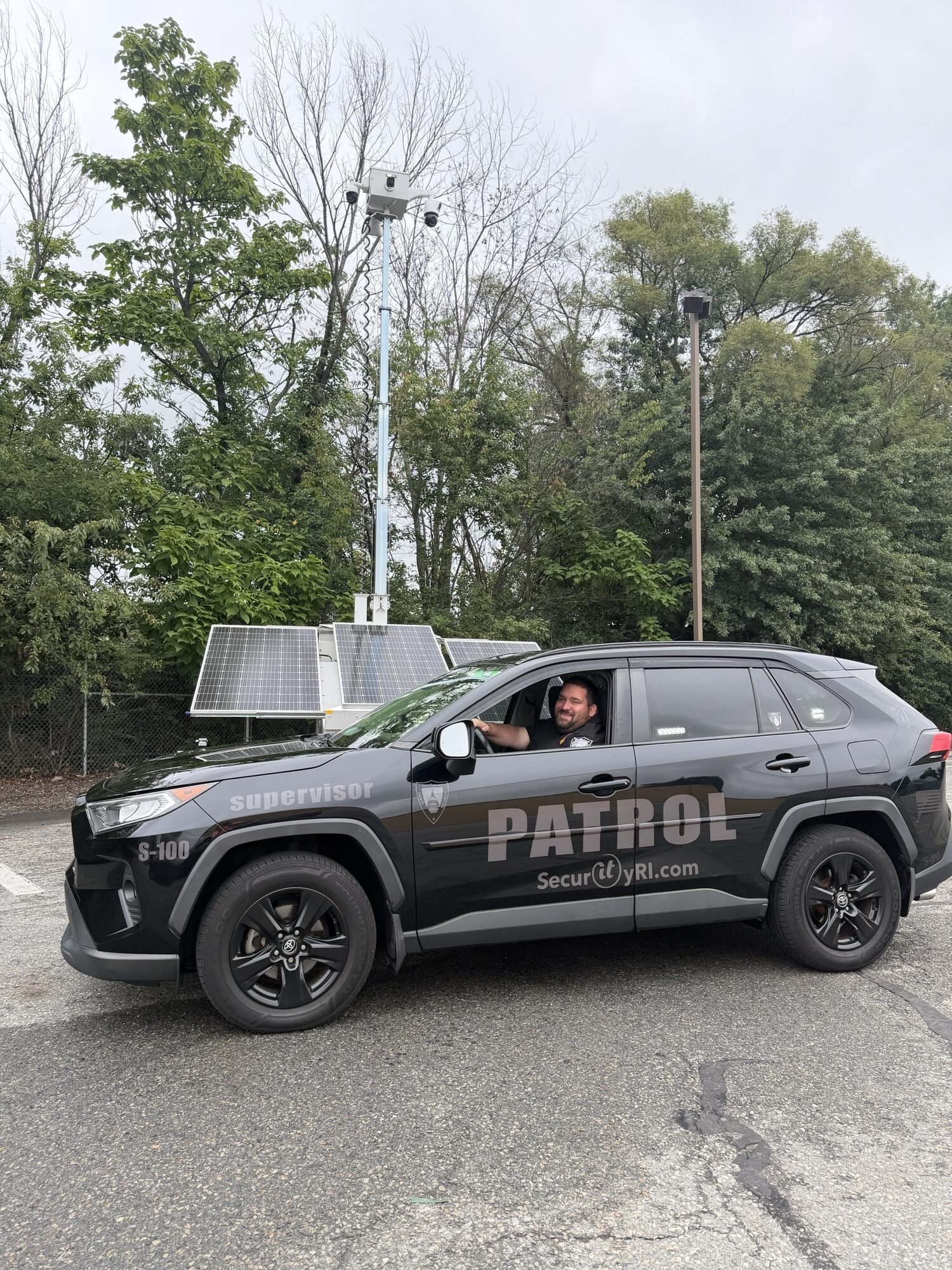 SecurityRI Lieutenant Vose patrolling a Rhode Island retail lot alongside a parked vehicle and mobile security trailer, tagging vehicles and enforcing lot rules